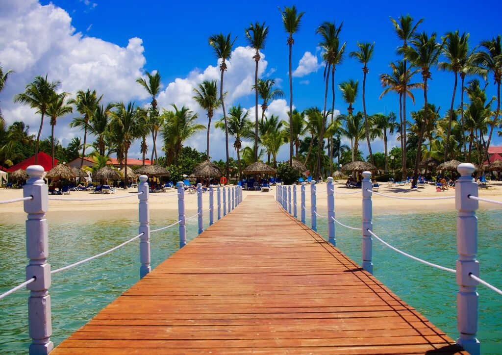 Wooden dock leading over clear turquoise Caribbean water to a beachfront tropical resort with palm trees