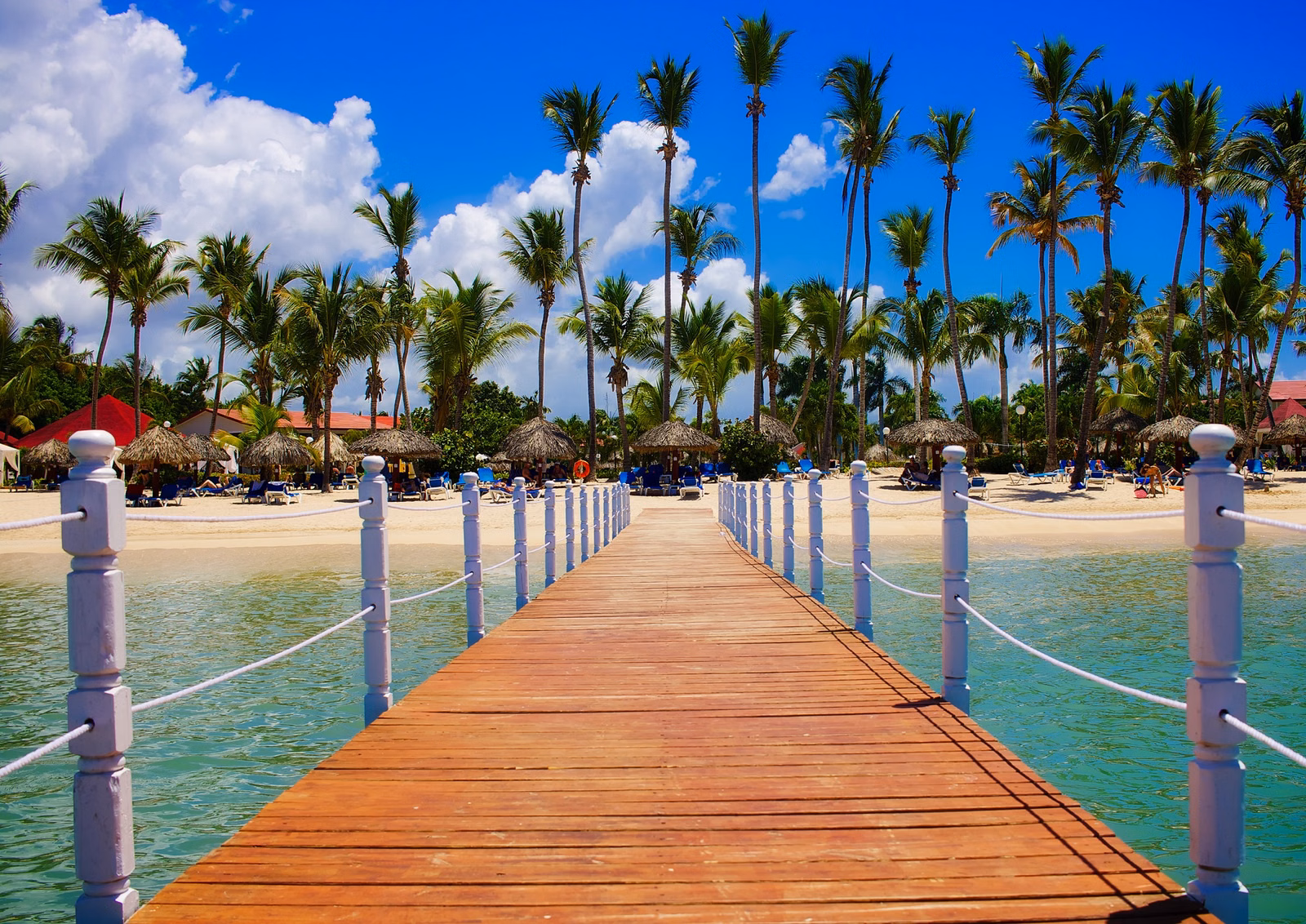 Wooden dock leading over clear turquoise Caribbean water to a beachfront tropical resort with palm trees