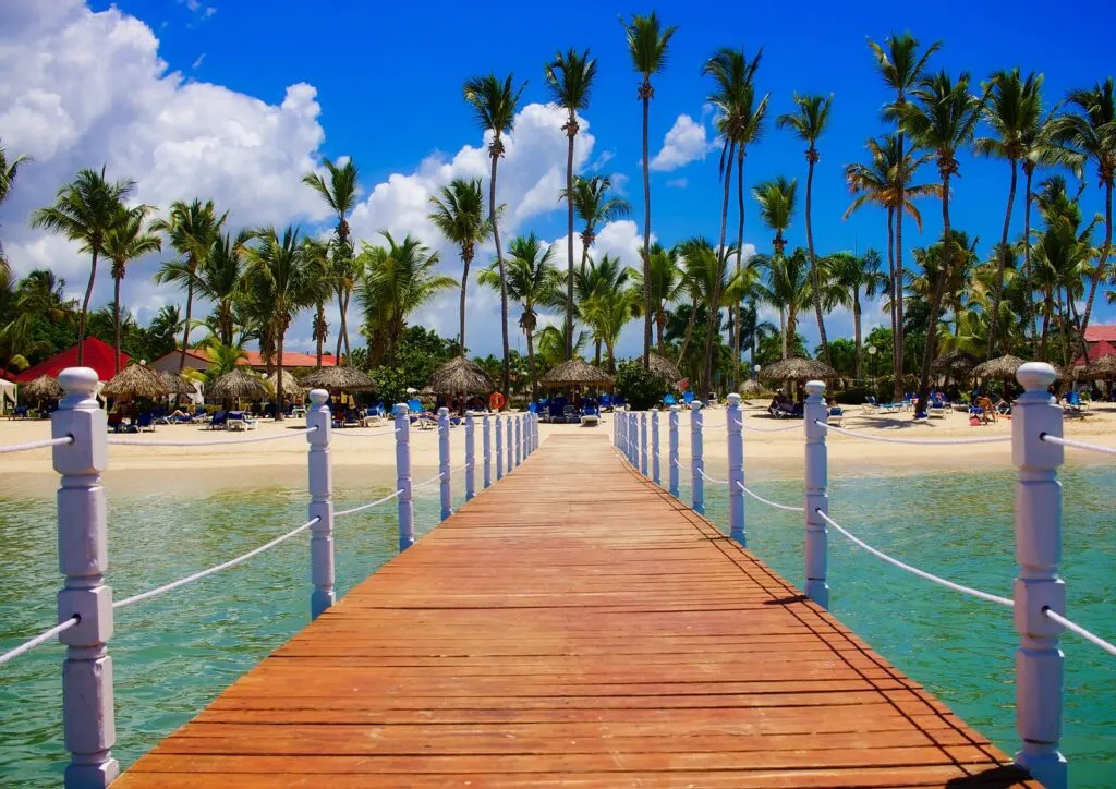 Wooden dock leading over clear turquoise Caribbean water to a beachfront tropical resort with palm trees